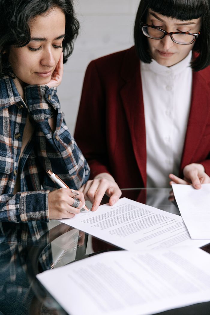 Home Two women reviewing and signing documents together at a glass table indoors.